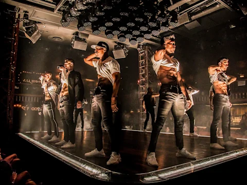A group of male dancers in white shirts and black pants perform on a stage under dramatic lighting in a nightclub setting.
