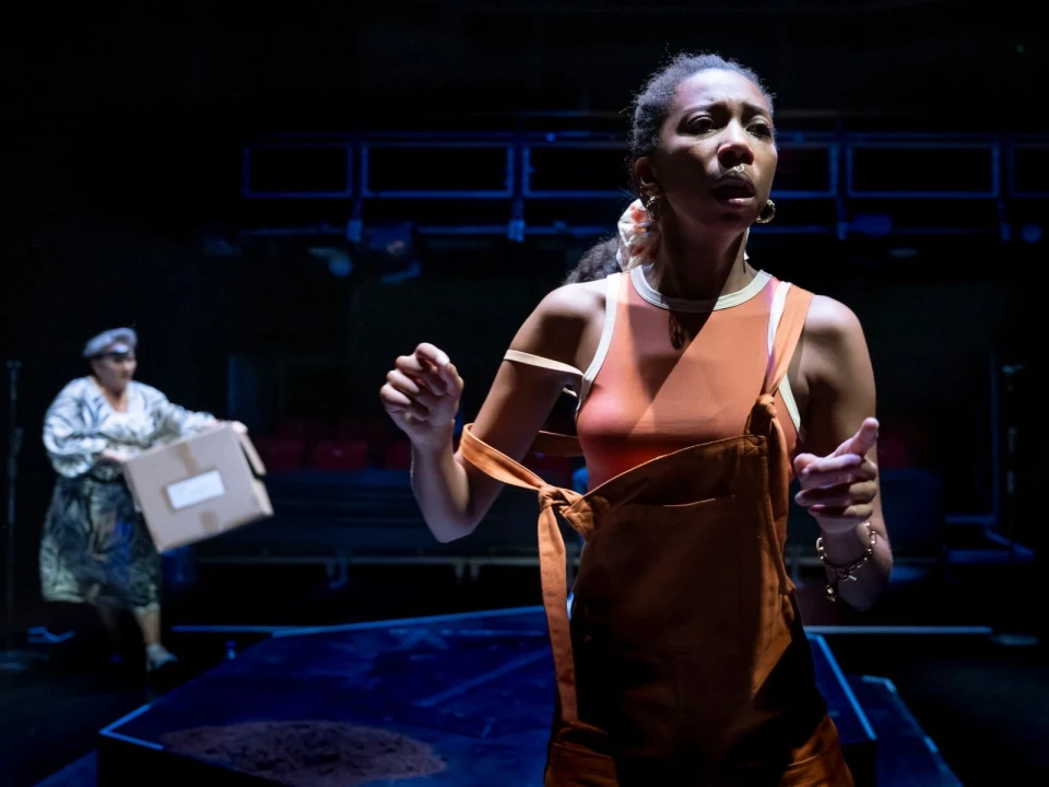 A woman in orange overalls stands on stage, looking focused. Another person is in the background, holding a cardboard box. The scene is dimly lit with a dark backdrop.