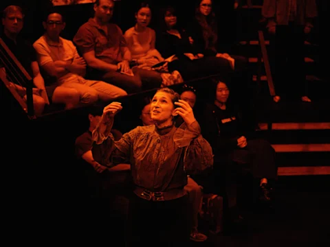 A performer gestures on stage under dramatic lighting while an audience watches from tiered seating in the background.