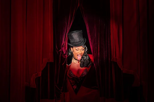 A person in a top hat and stage costume stands between red velvet curtains, sticking out their tongue and looking downward.