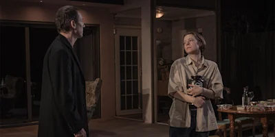 Two people stand in a dimly lit room; one faces the other, who is holding a large urn with both arms near a table set with food.