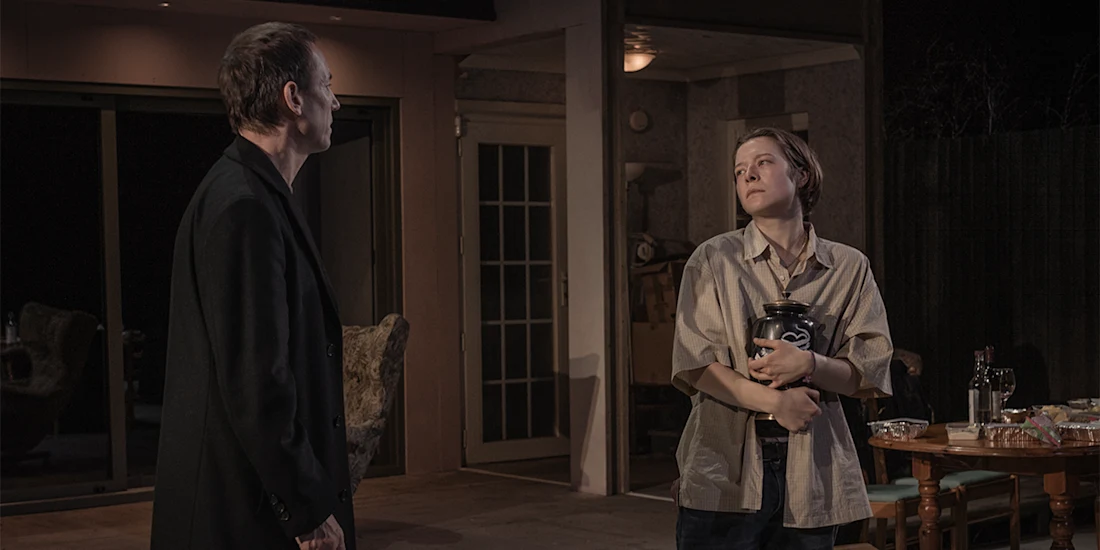 Two people stand in a dimly lit room; one faces the other, who is holding a large urn with both arms near a table set with food.
