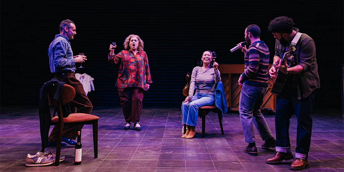 Five people perform on a stage; two are seated, three are standing, one holds a guitar, and another sings energetically. A wine bottle and glass are on the floor.