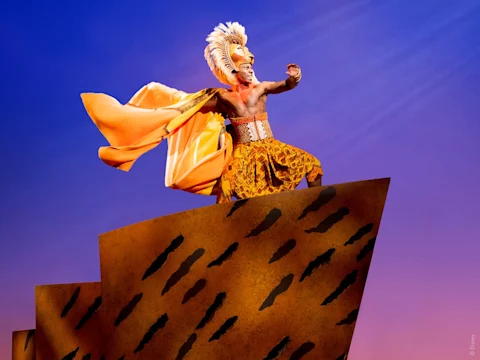 A performer in a lion costume stands on a stylized rock set against a purple sky, appearing to sing or gesture in a theatrical production.