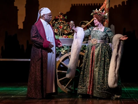 Two actors in elaborate costumes perform on stage; one is dressed as Scrooge in a nightcap and robe, the other as a festive figure in green with a jeweled crown and fur-trimmed cloak.
