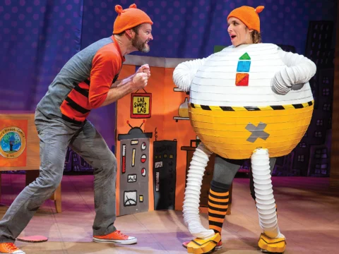 Two actors perform on stage. One actor wears a striped shirt and hat, while the other is dressed in a round, robotic costume. The stage backdrop features a cityscape and a colorful, whimsical setting.