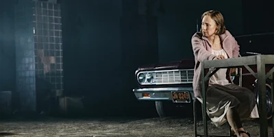 A woman in a light dress and pink coat sits at a metal table in front of a vintage red car in a dimly lit, industrial setting.
