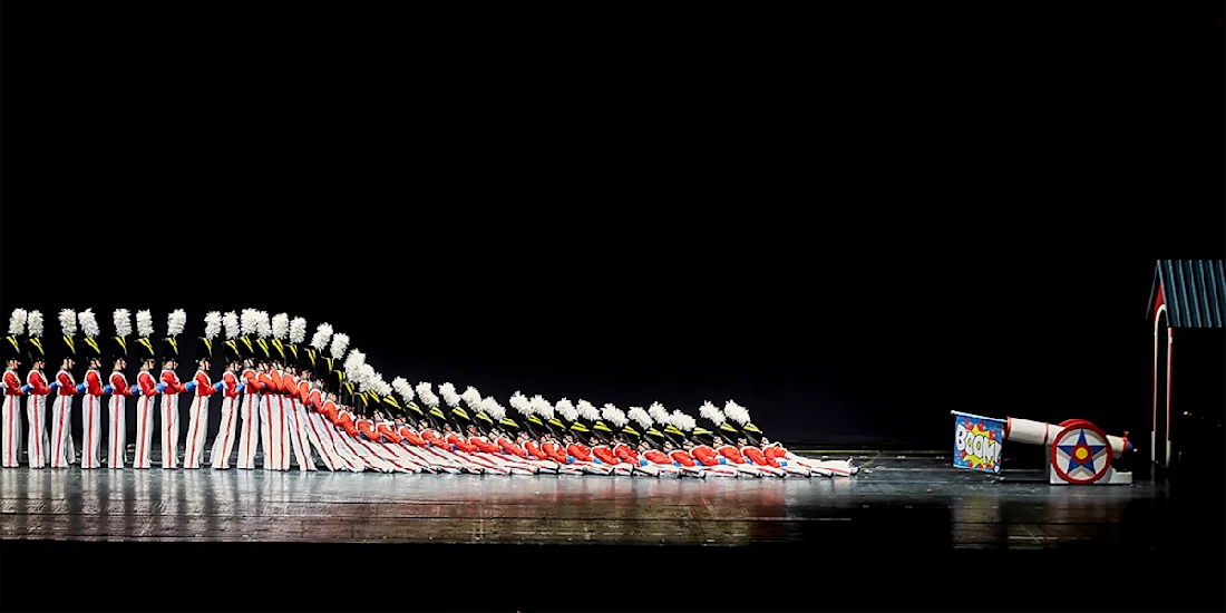 A line of performers dressed as toy soldiers fall in a domino effect on stage, with a toy cannon and small house structure at the end of the row.