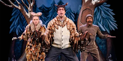 Three actors in animal costumes pose on a stage with a forest backdrop; one wears antlers, one has mouse ears, and one wears a brown outfit and hat.