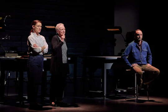 Three actors on stage, two standing and one sitting, perform in a dimly lit theatrical setting with tables and books in the background.