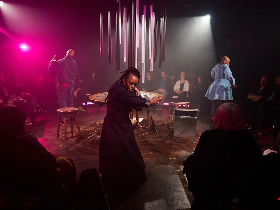 Three performers on a circular stage surrounded by an audience, with large metal chimes hanging above a table.