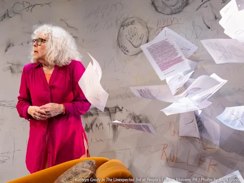 Kathryn Grody stands in front of a wall covered in writing and drawings while papers float through the air around her.