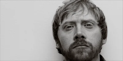 A close-up black and white photo of a light-skinned man with short hair and a beard, looking directly at the camera with a neutral expression.