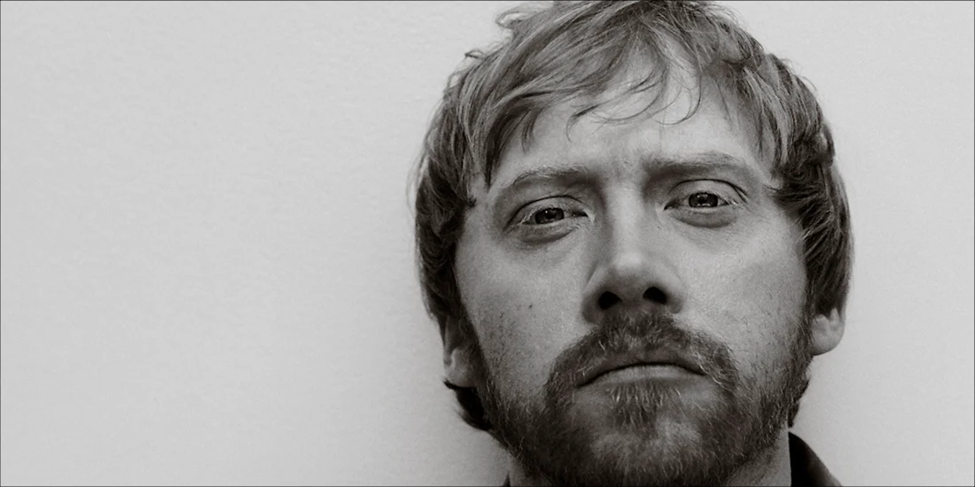 A close-up black and white photo of a light-skinned man with short hair and a beard, looking directly at the camera with a neutral expression.