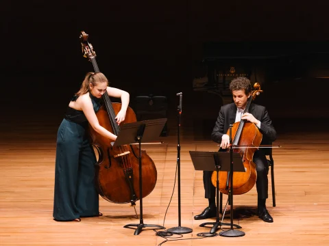 A woman playing a double bass and a man playing a cello perform on a stage, both standing next to music stands. A grand piano is in the background.