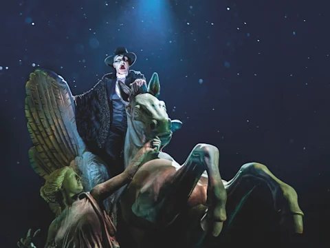 A person in theatrical makeup and costume sits atop a winged horse statue under dramatic lighting, with a female statue figure in the foreground.