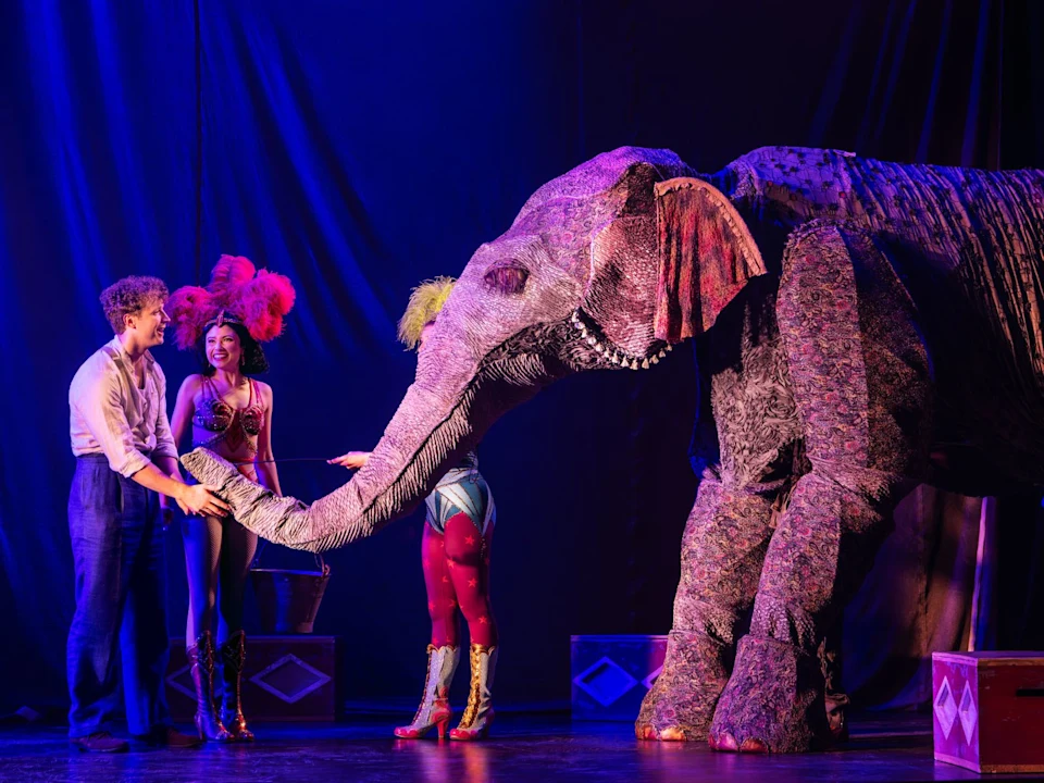 Two performers stand with a person in a fabric elephant costume on stage, with colorful lighting and circus-themed props in the background.