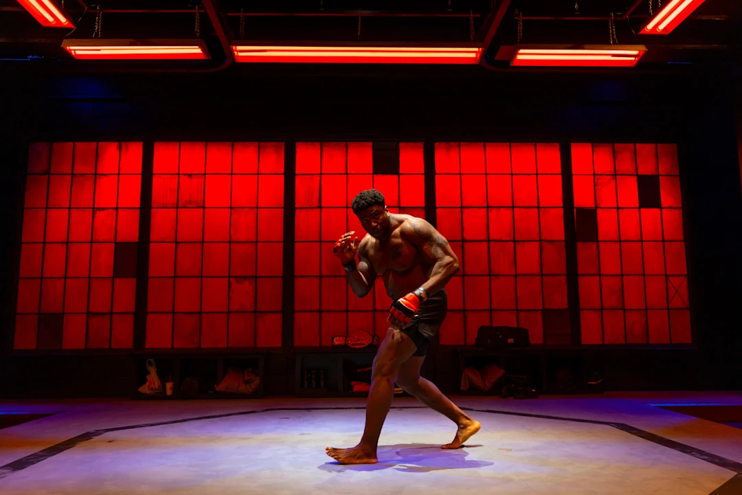 A shirtless male mixed martial artist in fight stance stands on an octagon mat, illuminated by red lighting, with boxing gloves and gear in the background.