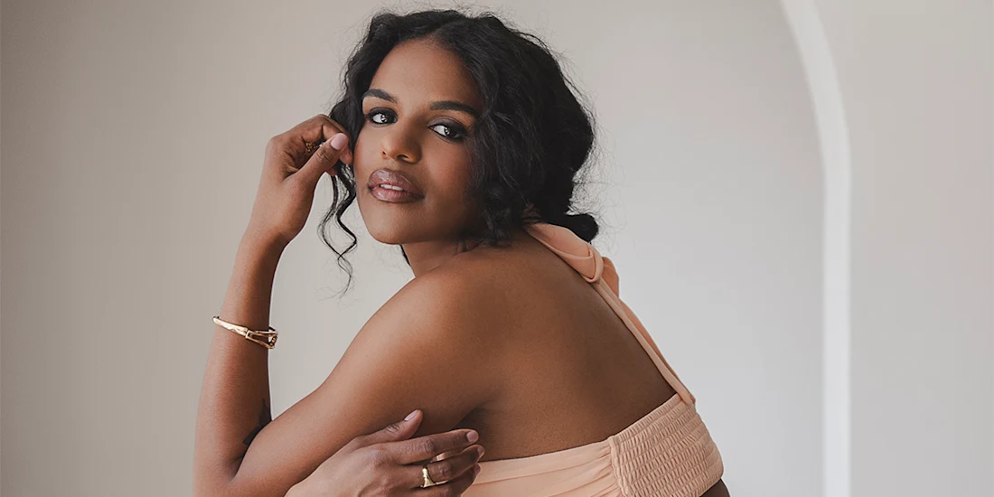 Woman in a peach sleeveless dress sits sideways, looking over her shoulder, with one hand touching her face and the other resting on her arm.