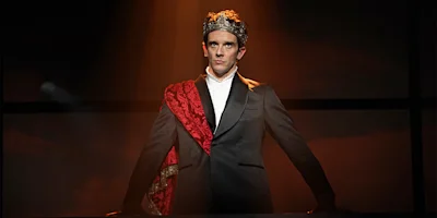 A man in a suit wears a crown and red sash, standing under a spotlight on stage with a serious expression.