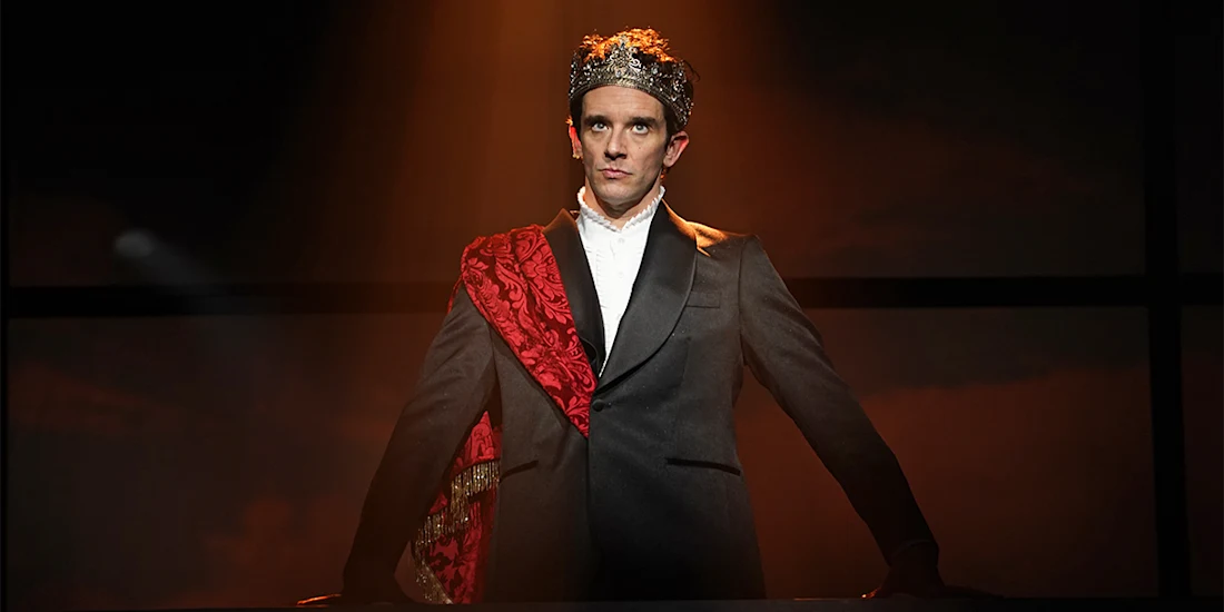 A man in a suit wears a crown and red sash, standing under a spotlight on stage with a serious expression.