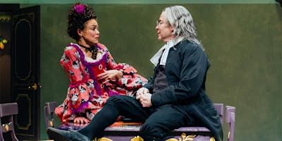Two actors in period costumes sit on a table during a stage performance, engaged in an animated conversation against a simple set background.