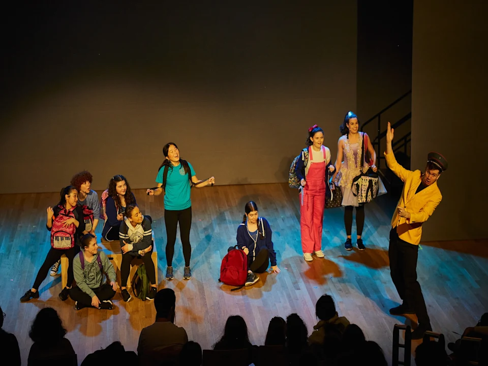 A group of performers on stage, with nine people sitting and standing, and one in a yellow coat gesturing.