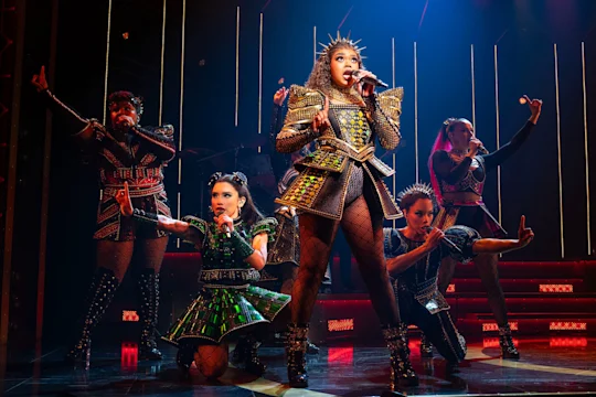 Six performers in ornate, metallic costumes sing and pose on stage with dramatic lighting and illuminated vertical lines in the background.