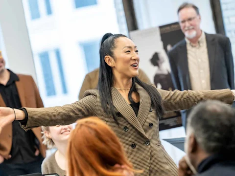 A woman in a brown plaid blazer stands with arms outstretched, smiling and speaking to a group of people in a bright room.