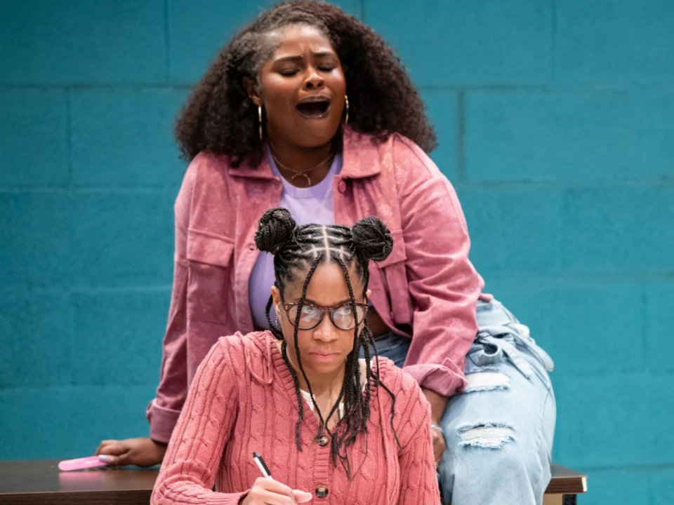 Two women indoors; one with braids and glasses writing while sitting at a table, and the other with curly hair and earrings sitting on the table behind her, appearing to be singing or speaking.