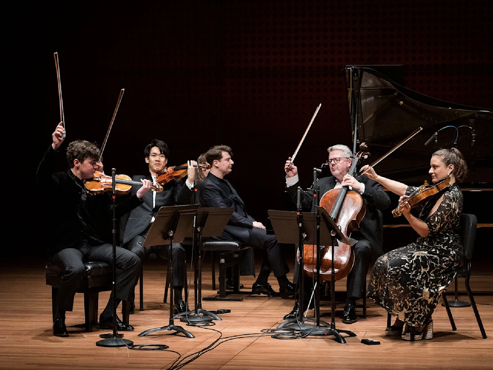 Five musicians with string instruments and a grand piano sit on stage, holding up their bows, appearing to acknowledge applause after a performance.