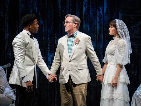 Three people stand on stage in costume: one in a white tuxedo, one in a suit with a bow tie holding both hands, and one in a wedding dress against a dark backdrop.