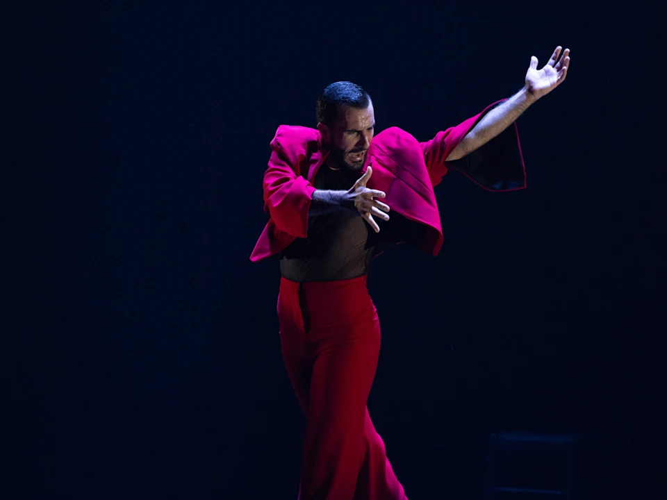 A person in a red outfit performs a dramatic dance pose on a dimly lit stage, with one arm extended and expressive facial expression.