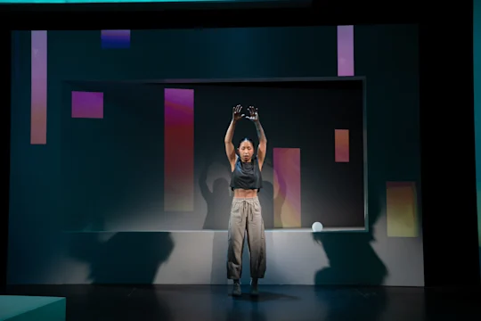 A person stands on stage with arms raised, wearing a black crop top and gray pants, in front of a geometric background with colorful rectangular lights.