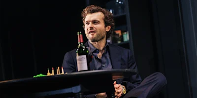 A man in a dark suit sits at a table with a wine bottle and snacks, looking to the side with a relaxed expression in a dimly lit room.