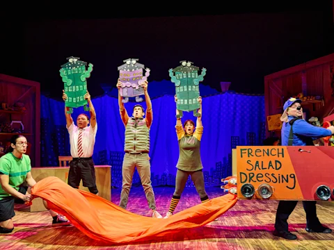 Five actors perform on stage with colorful props, including building cutouts and a cardboard car labeled "French Salad Dressing," on a brightly lit theatrical set.