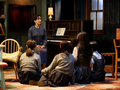 A woman sits near a piano facing five children sitting on the floor, all indoors in a warmly lit room with wooden furniture.