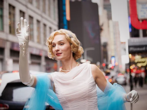 Woman in a white dress and blue shawl waving on a city street.