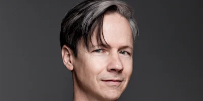 A man with short, straight, dark and gray hair is facing the camera and smiling slightly against a plain gray background.