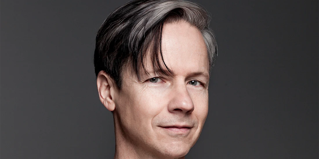 A man with short, straight, dark and gray hair is facing the camera and smiling slightly against a plain gray background.