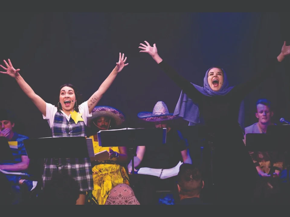 Two performers stand with arms raised and mouths open on stage in front of music stands, while others in hats sit and read scripts behind them.