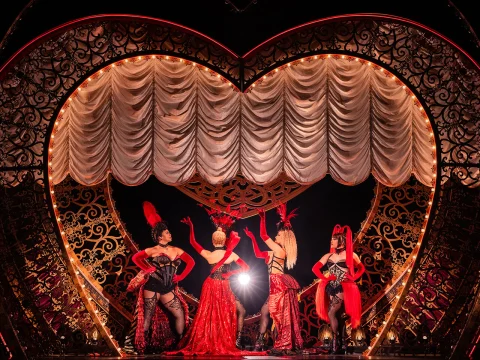 Performers in elaborate red and black costumes stand on stage in front of a heart-shaped backdrop with ornate details and draped curtains.