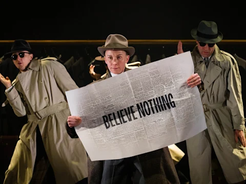 Three people in trench coats and hats stand on stage; the center person holds a large newspaper with “BELIEVE NOTHING” printed in bold letters across the front.