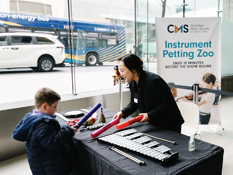 A woman helps a young boy try percussion instruments at an "Instrument Petting Zoo" table inside a building, with a city bus and street visible through the window.