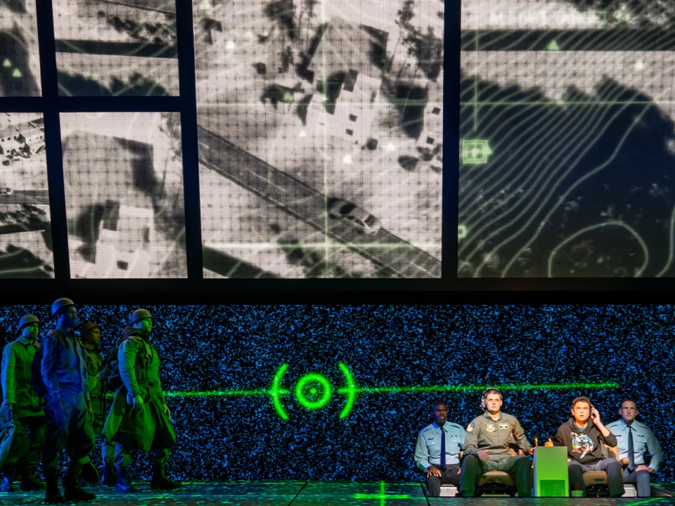 A group of people in military uniforms sit facing a large screen displaying an aerial map with a green target reticle, while other uniformed individuals stand nearby.