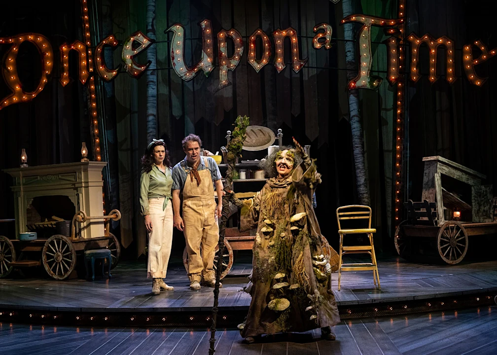 Three actors perform onstage; two stand together while one, dressed as a tree, gestures excitedly. The set features vintage furniture and a large marquee sign reading “Once Upon a Time.”.