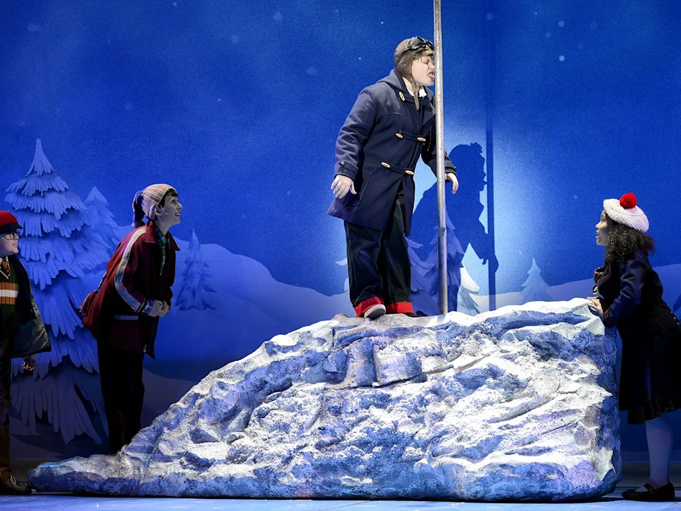 Four children in winter clothing perform a scene on a stage set with a snowy hill and a metal pole.