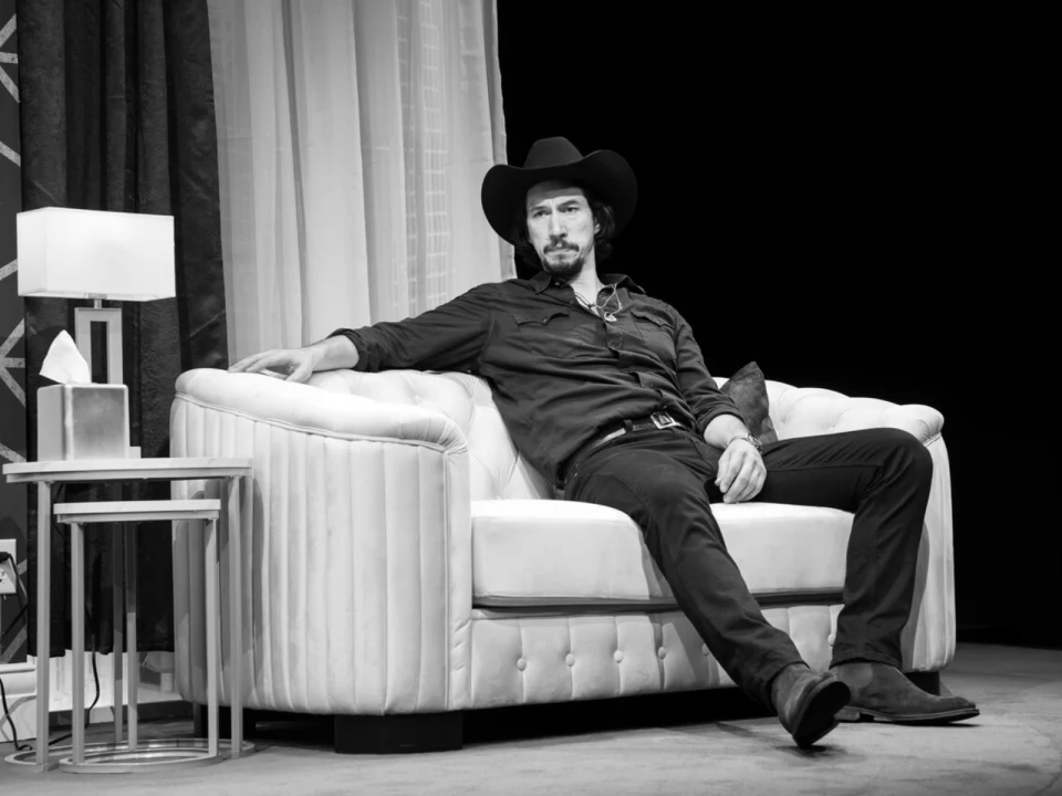 A person wearing a cowboy hat sits on a couch, arm resting on the back. The room features a lamp and drapes. Image is in black and white.