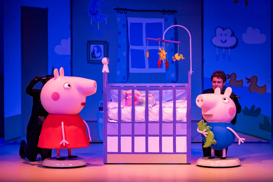 Two performers operate Peppa Pig and George puppets next to a crib on a brightly lit stage with a baby mobile and cartoon-style nursery backdrop.