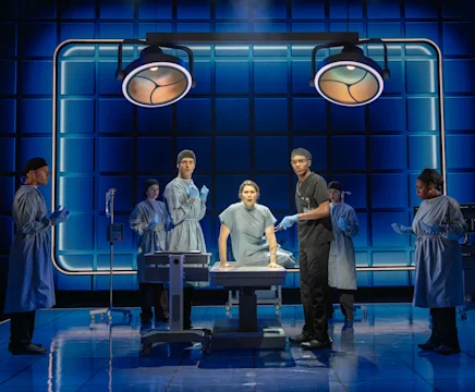 A patient sits on an operating table surrounded by six medical professionals in a blue-lit theater set.